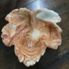 A ceramic dish shaped like a leaf with wavy, uneven edges sits on a dark wooden surface. The dish features a marbled design in shades of orange, cream, and light brown, mimicking natural leaf patterns. Light reflects off its glossy surface.