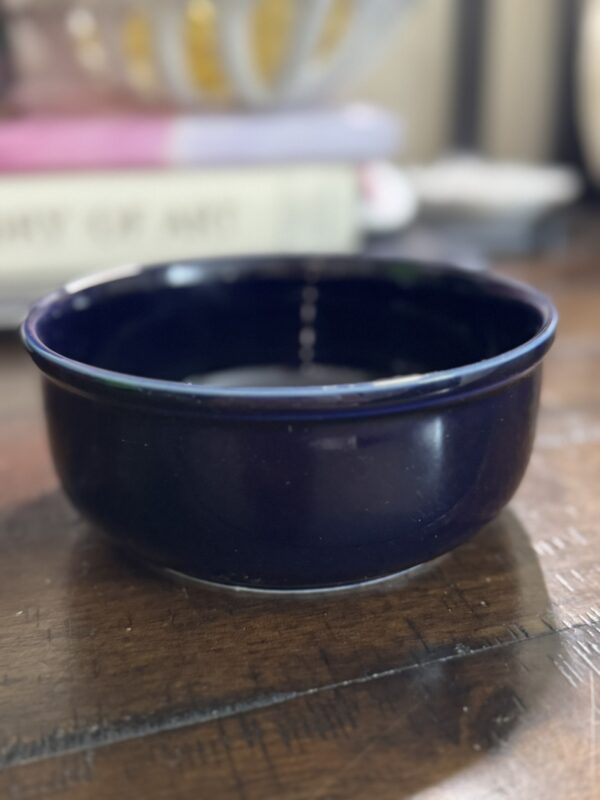 A deep, dark blue ceramic bowl sits on a wooden surface. The background is blurred, showing the spine of a closed book and part of a yellow object, possibly a fruit basket. The lighting is soft and natural.