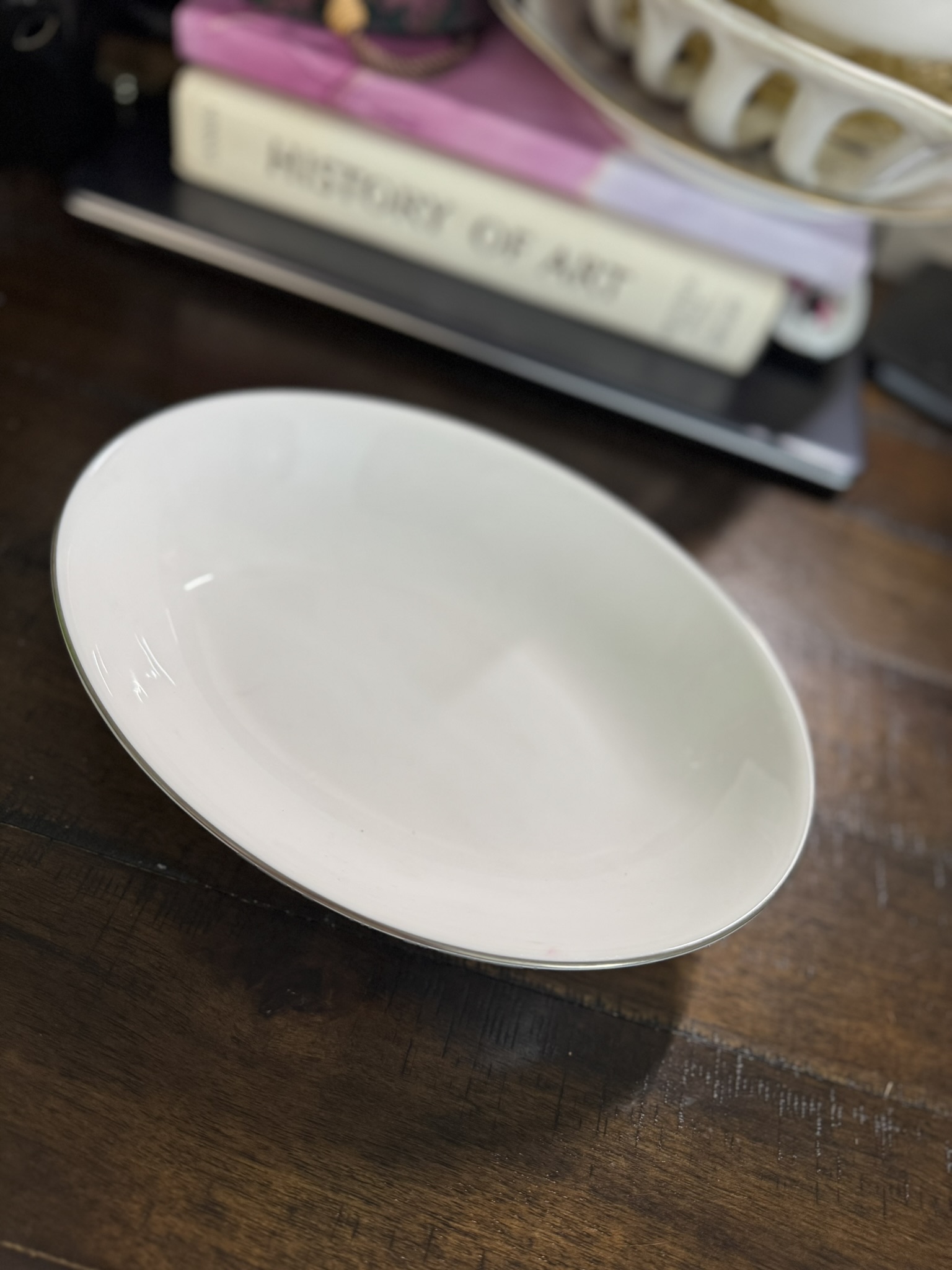 A white, oval-shaped ceramic bowl sits on a dark wooden table. In the blurred background, there is a stack of books, with one titled HISTORY OF ART visible, and a white decorative object resting on top of the books.
