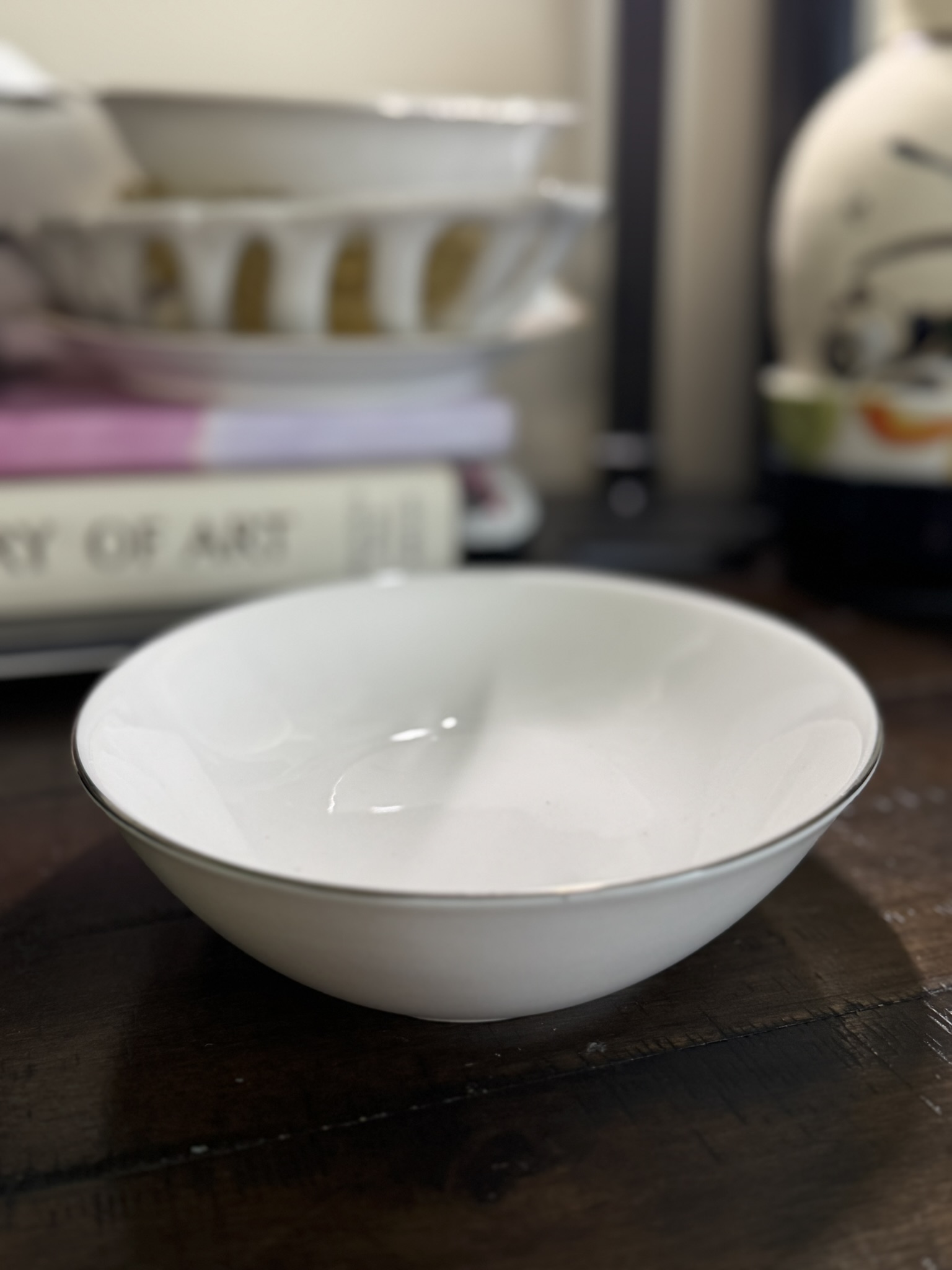 A white ceramic bowl rests on a dark wooden surface, while the Blue Crackle Glass Vase, stacked dishes, and the book “ART OF ART” create a softly lit, atmospheric backdrop.