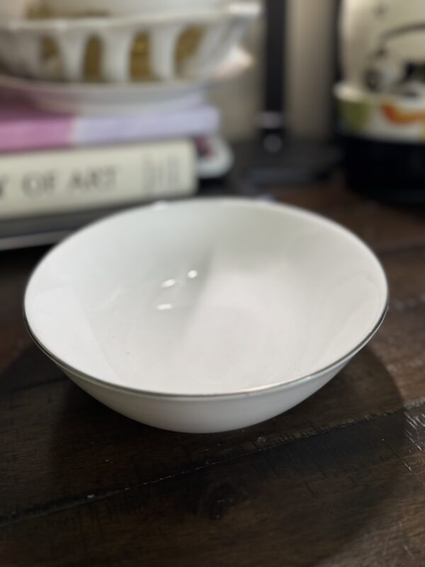 A plain white ceramic bowl rests on a dark wooden table. In the background is a Blue Crackle Glass Vase, stacked books (one titled OF ART), and softly blurred items like a plate, cup, and other household objects.