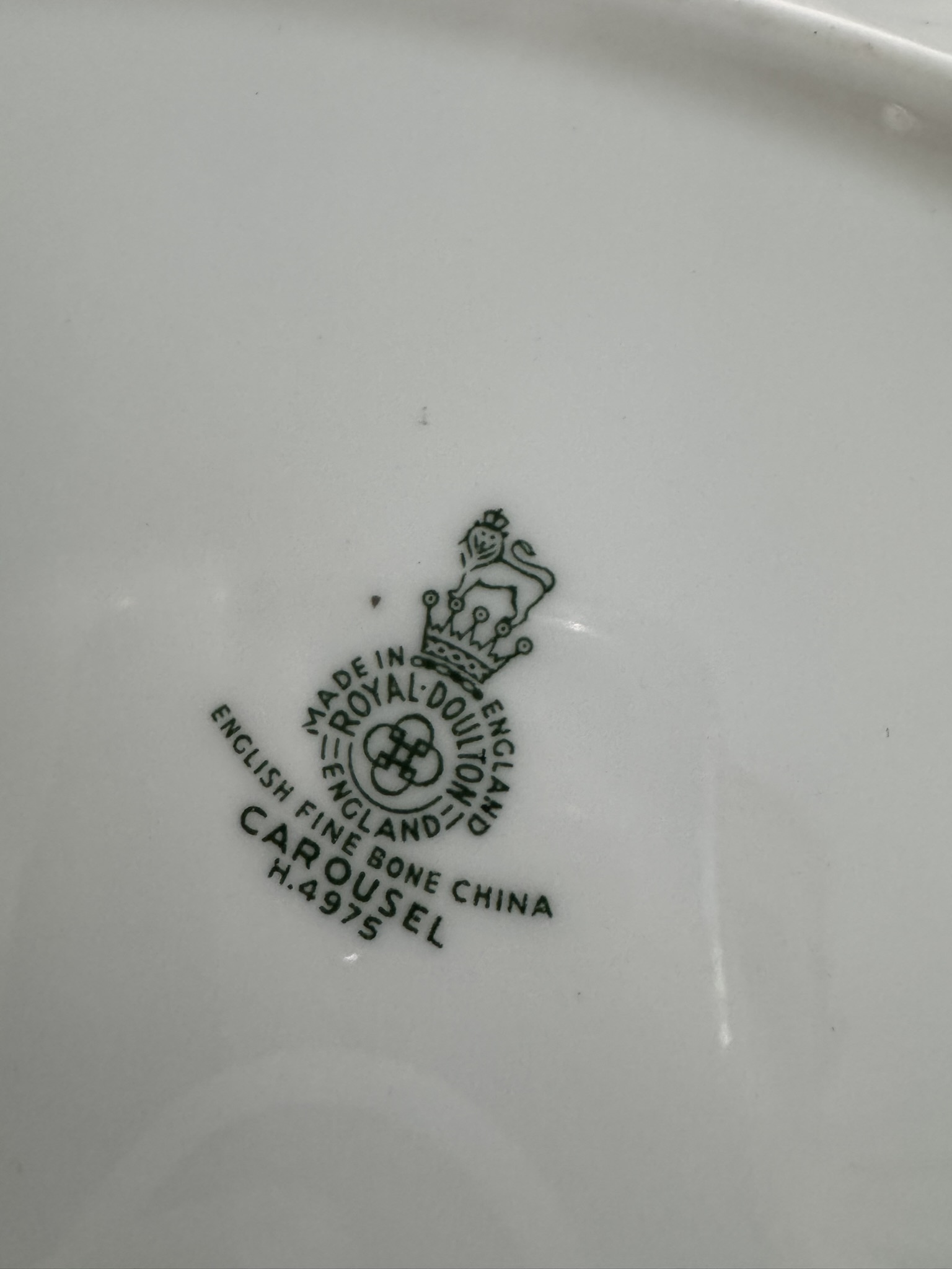 Close-up of a ceramic plates underside showing a green Royal Doulton England hallmark. The logo includes a lion and crown, and text reads: “MADE IN ENGLAND, ROYAL DOULTON, ENGLAND, ENGLISH FINE BONE CHINA, CAROUSEL, H.4915.”.