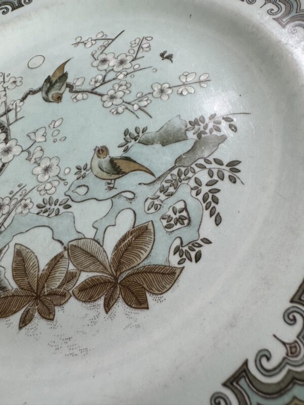 A close-up of a decorative ceramic plate featuring two small birds perched on blossoming branches, with brown leaves and intricate floral patterns in soft blues and browns. The plate’s ornate border is visible along the edge.