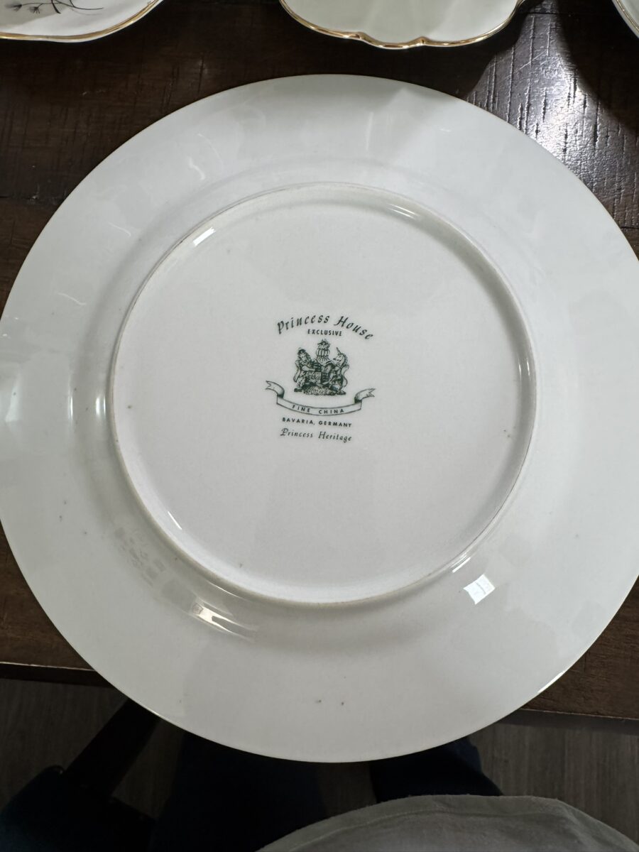 The underside of a white ceramic plate, showing the maker’s mark in green. The mark reads Princess House Exclusive, Bavaria Germany, Princess Heritage with a crest and two lions in the center. The plate rests on a dark wooden surface.