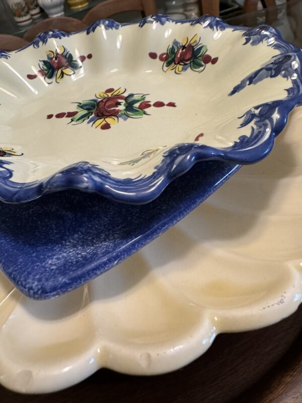 Three decorative ceramic plates are stacked. The top plate is cream with blue edges and floral patterns. The middle plate is square and speckled blue. The bottom plate is round, cream-colored, and scalloped, all sitting on a wooden surface.