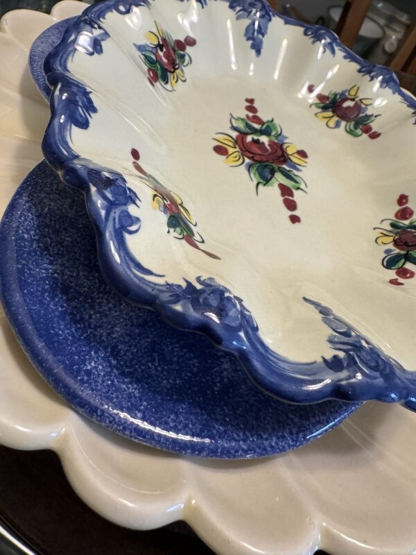 Three decorative ceramic plates are stacked. The top plate is cream-colored with blue edges and floral designs in red, yellow, and green. The middle plate is blue with a textured pattern. The bottom plate is cream with a scalloped edge.