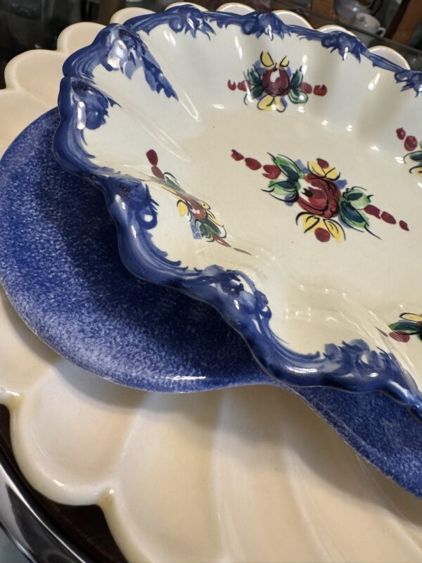 Three ceramic plates are stacked: a cream-colored plate with scalloped edges at the bottom, a speckled blue plate in the middle, and on top, a cream plate with blue scalloped trim and a hand-painted red, yellow, and green floral design in the center.