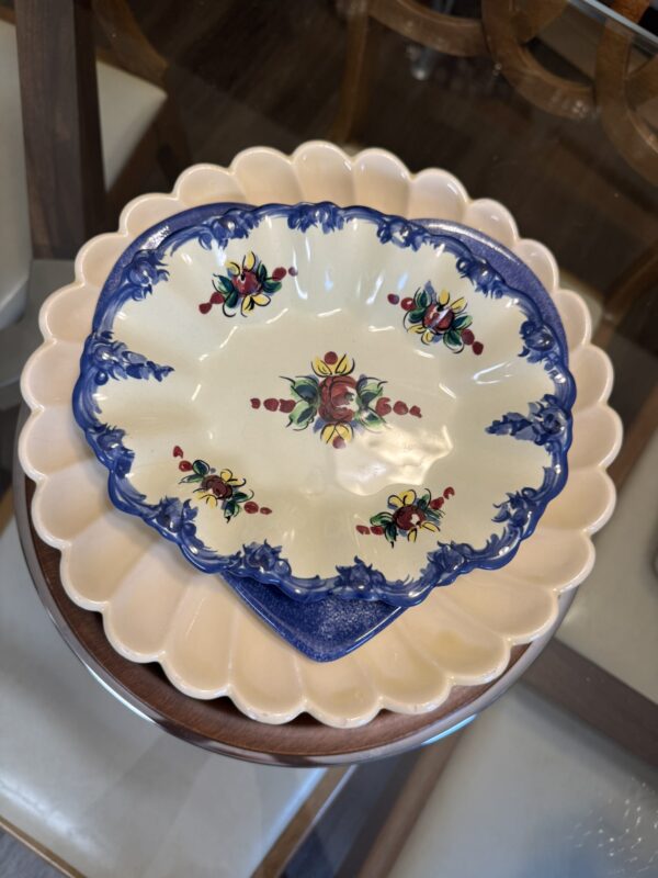A scalloped ceramic plate with a cream center, blue edges, and floral designs overlaps a larger, pale pink, scallop-edged plate, both set on a glass table with wooden edges and cream-colored cushioned chairs visible around it.
