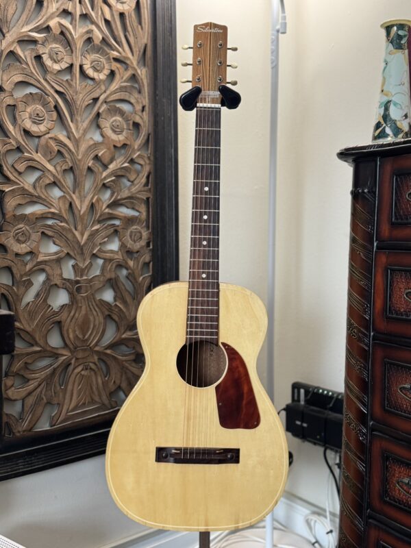 A light-colored Silvertone 1960's 699 Guitar with a dark wood neck and headstock stands on a black stand near an ornate carved wood panel, a brown chest of drawers with a lamp, and a ceramic vase.
