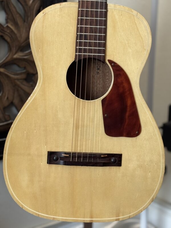 A close-up of the Silvertone 1960's 699 Guitar’s body highlights its light wood finish, dark wood bridge, reddish-brown pickguard, six steel strings, soundhole, and fretted neck set against blurred ornate wood decor in the background.