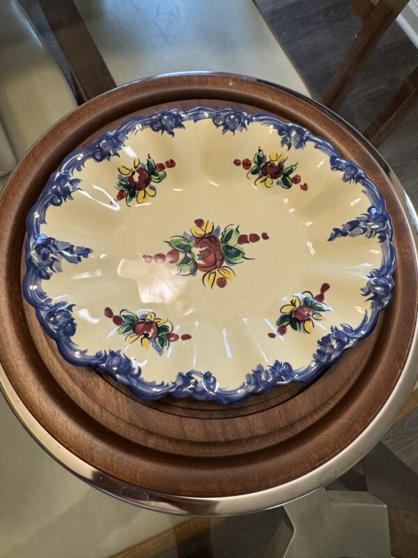 Ornate ceramic platter with blue scalloped edges and colorful hand-painted floral designs, featuring red, yellow, and green accents. The platter rests on a round wooden tray, both on a glass tabletop with visible furniture legs beneath.