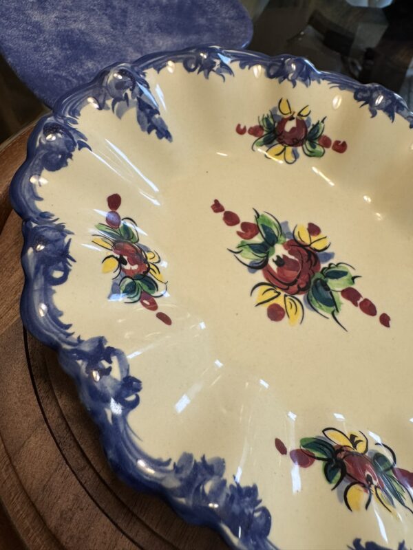 A cream-colored ceramic plate with scalloped edges and blue floral trim sits atop a wooden surface. The plate is decorated with painted red and yellow flowers with green leaves and dotted red accents in its center and around the rim.