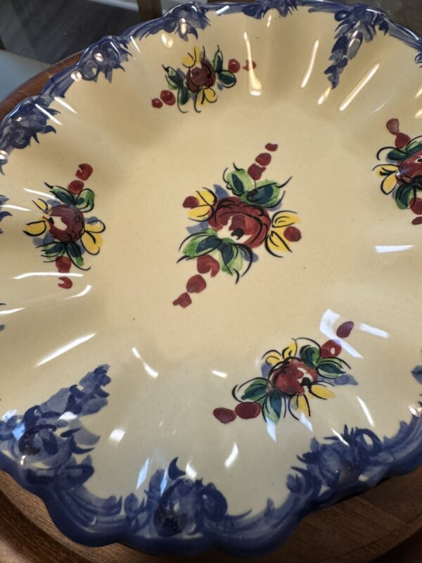 A ceramic plate with scalloped edges, featuring blue painted trim and floral patterns in red, yellow, and green. The plate has a cream background and is displayed on a wooden surface. The floral design appears three times around the rim and once in the center.