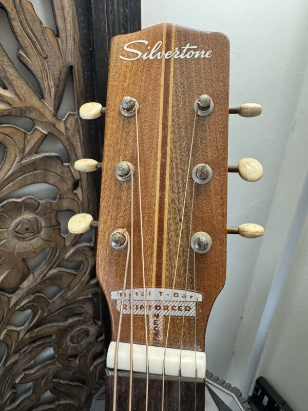 Close-up of a Silvertone 1960's 699 Guitar headstock with six cream tuners, striped wood center, and cursive Silvertone logo. An ornate carved wooden panel appears in the background.