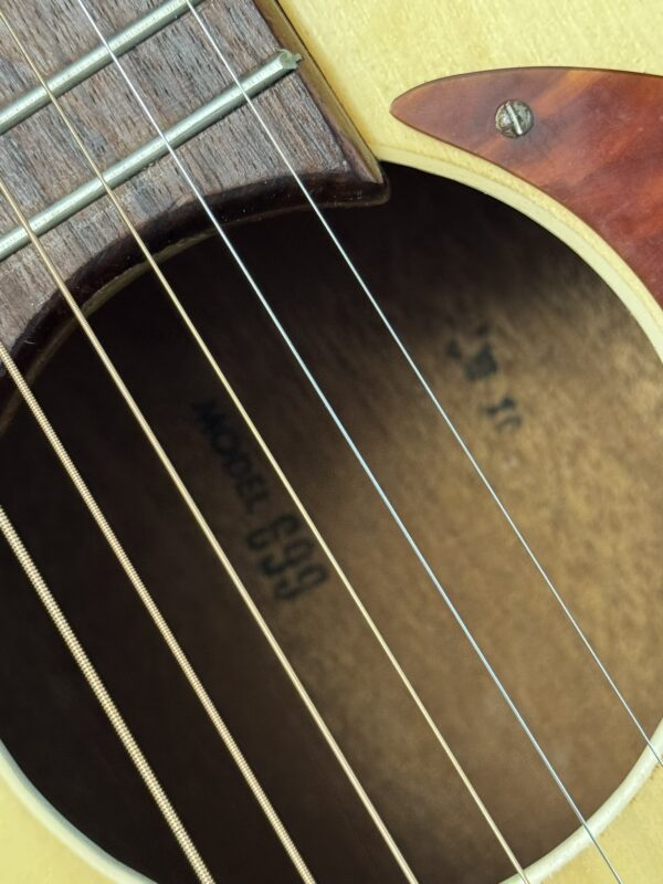 Close-up of the Silvertone 1960's 699 Guitar sound hole reveals metal strings, part of the wooden bridge, and a “MODEL 699” label. The wood grain and pickguard highlight this classic guitar’s vintage craftsmanship.