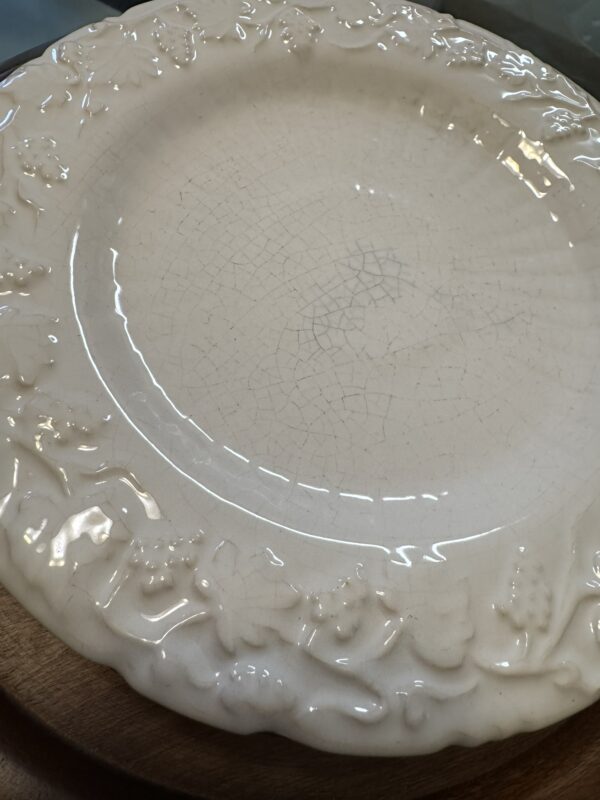 A close-up of a cream-colored ceramic plate with a crackled glaze. The rim is decorated with a raised floral and grapevine pattern. The plate rests on a wooden surface, and light reflects off its glossy finish.