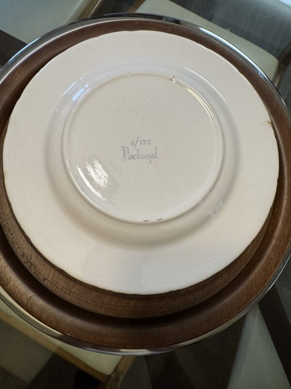 A white, vintage ceramic plate with visible crackling on its underside rests on a dark wooden plate. The plate is marked “4/173 Portugal” in blue lettering at the center. Both plates are on a glass table with cream-colored chairs visible around it.