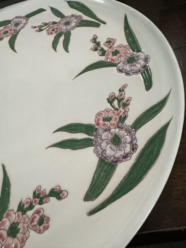 A round white ceramic plate with hand-painted floral designs. The flowers have pink and purple petals with green leaves, arranged along the rim of the plate. The plate rests on a dark wooden surface.