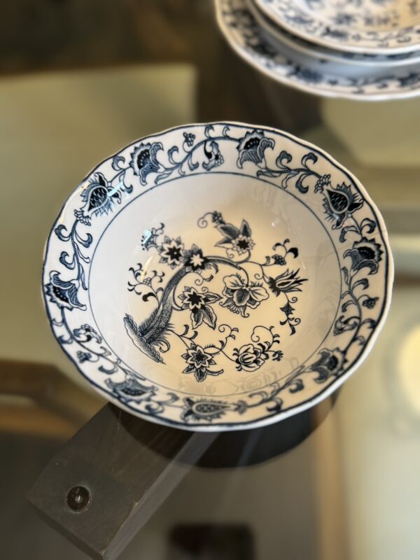 A white ceramic bowl with intricate blue floral and bird patterns on the inner and outer edges sits on a glass table. The design includes swirling vines and flowers. Other matching plates are partially visible in the blurred background.