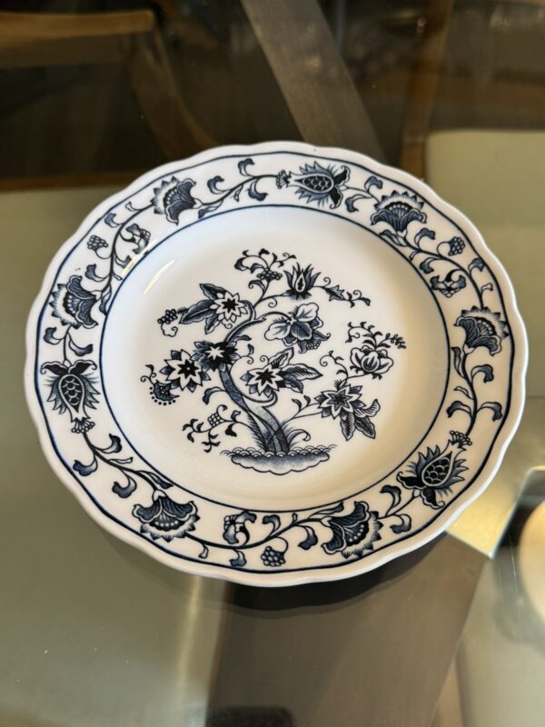 A white ceramic plate with a blue floral and leaf design along the scalloped rim and a central blue floral motif, resting on a glass table with brown wooden chairs visible in the blurred background.