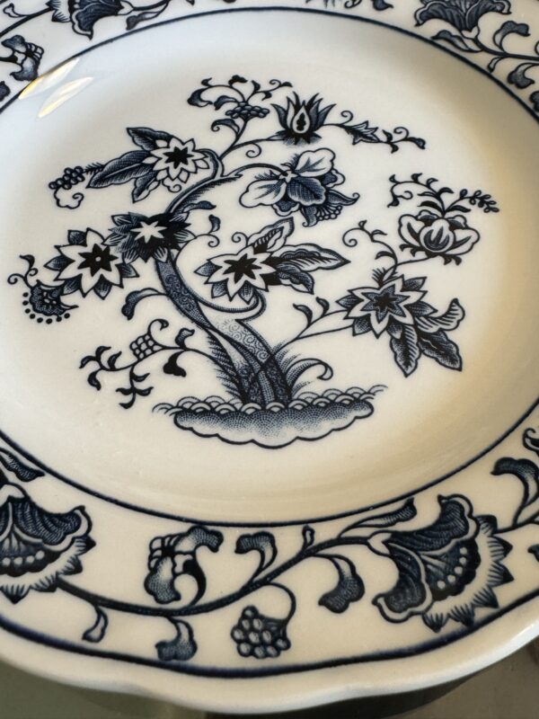 A close-up of a white ceramic plate featuring intricate blue floral patterns. The plate’s center showcases a stylized tree with detailed flowers and leaves, while the border displays more ornate floral and leafy designs, all in dark blue ink.