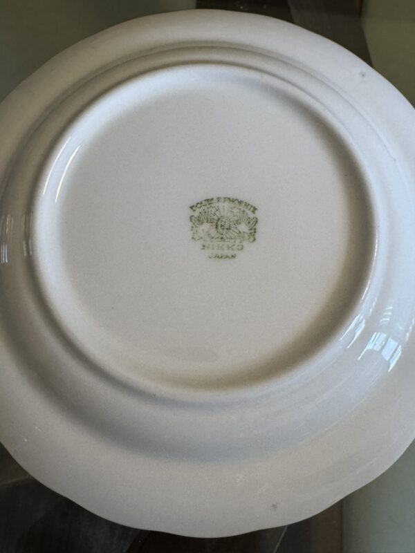 The image shows the underside of a white porcelain plate with a green stamp that reads Colle et Pissetti, Nikko, Japan, indicating its manufacturer and origin. The plate has a slightly scalloped edge and rests on a glass surface.