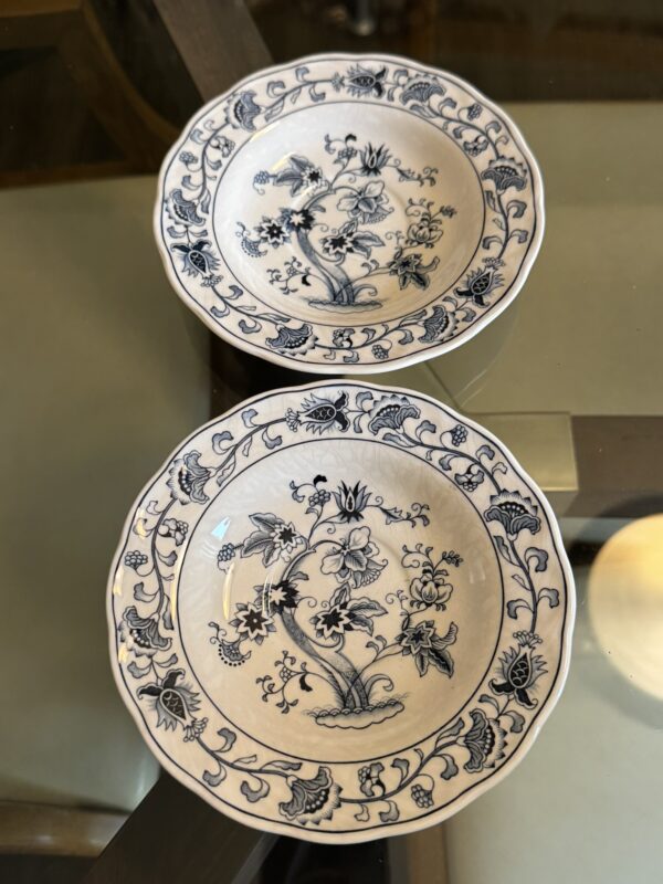 Two porcelain bowls with blue floral patterns sit on a glass table. The bowls have scalloped edges and feature intricate designs of flowers and leaves. The reflection of the table and part of a round white object are visible in the lower right corner.