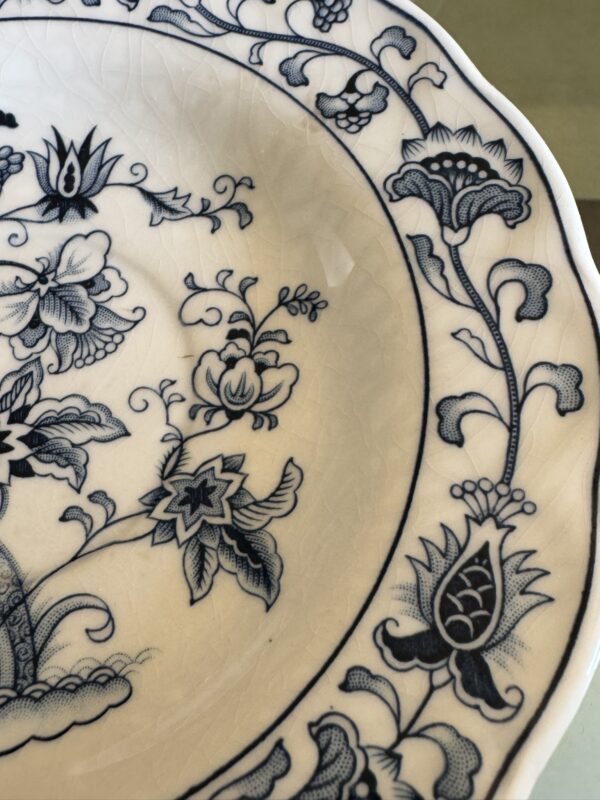 A close-up of a white ceramic plate with intricate blue floral and leaf patterns along the edge. The surface shows visible crackling, and the rim has a gently scalloped shape. The design features stylized flowers and vines in a traditional style.