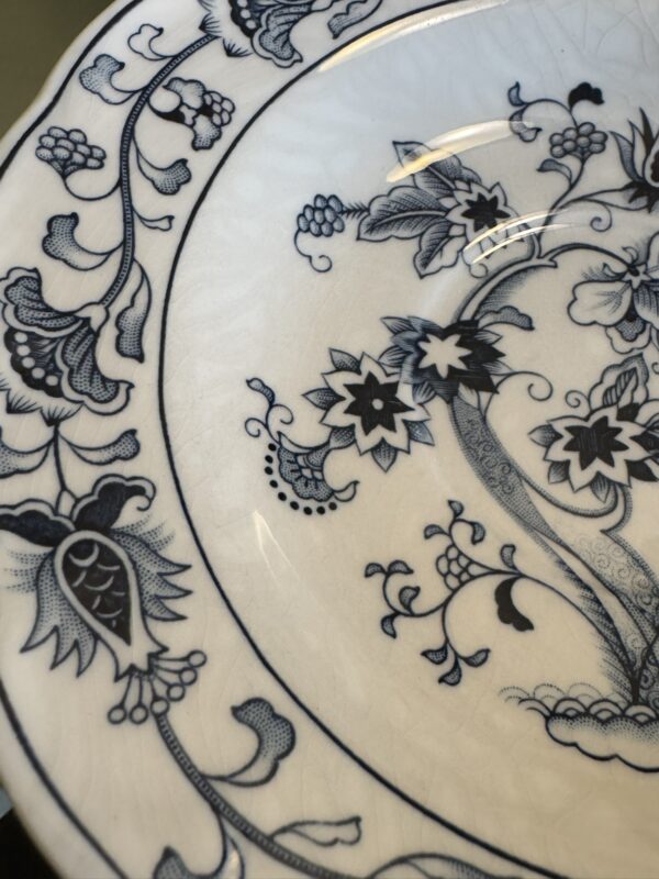 A close-up of a white ceramic plate with a blue floral pattern. The intricate design features stylized flowers, leaves, and vines, with fine detailing along the edge and surface. The plate shows faint crackling in the glaze, adding texture and character.