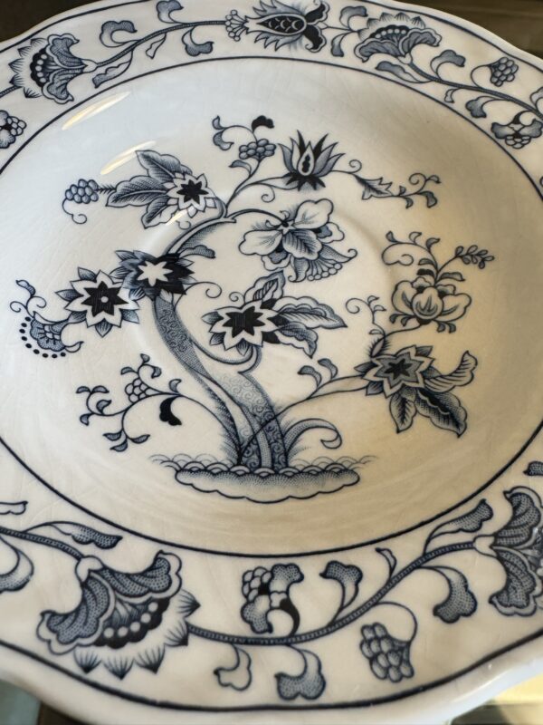 A ceramic plate with intricate blue floral and vine patterns on a cream background. The central design features stylized flowers with detailed leaves, surrounded by a decorative border of blooms, leaves, and abstract shapes. The plate has a glossy finish.