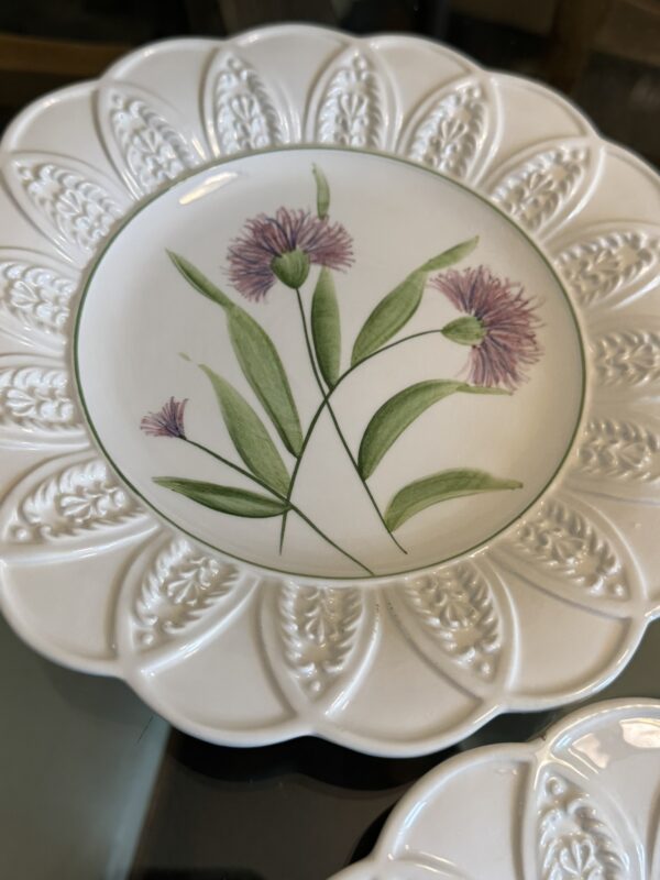 A decorative white ceramic plate with an embossed scalloped rim features a central design of purple and pink wildflowers with green leaves, set against a plain white background and bordered by a thin green line.