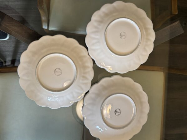 Three white, scalloped-edged plates are placed face down on a glass table, forming a loose triangle. Each plate shows a circular manufacturer’s mark on the bottom. Parts of wooden chair arms and cream-colored seat cushions are visible in the background.