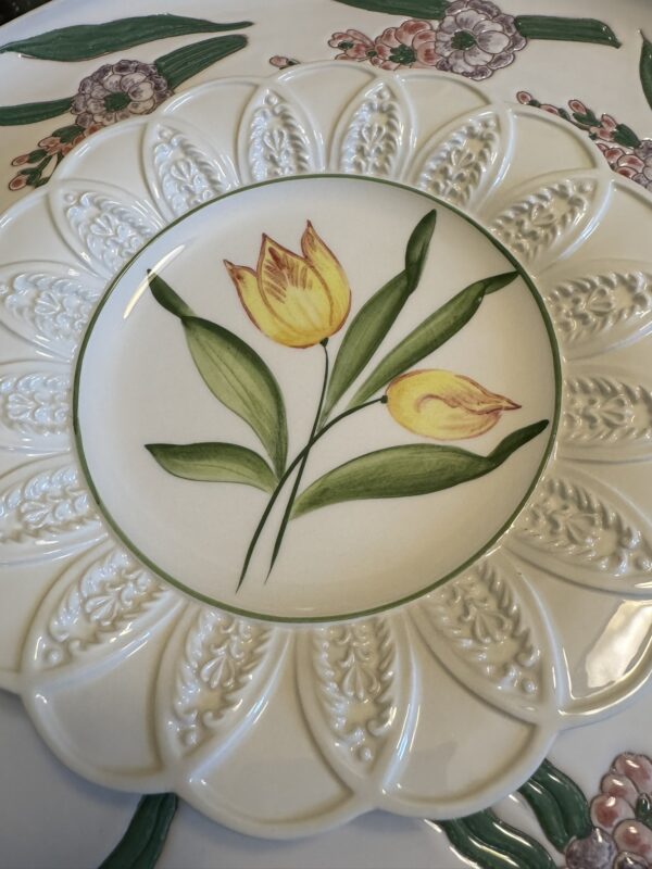 A decorative plate features two yellow tulips with green stems and leaves painted at the center. The plate has a textured, scalloped edge with a floral pattern, and rests on a larger dish with pink, green, and purple flower designs.