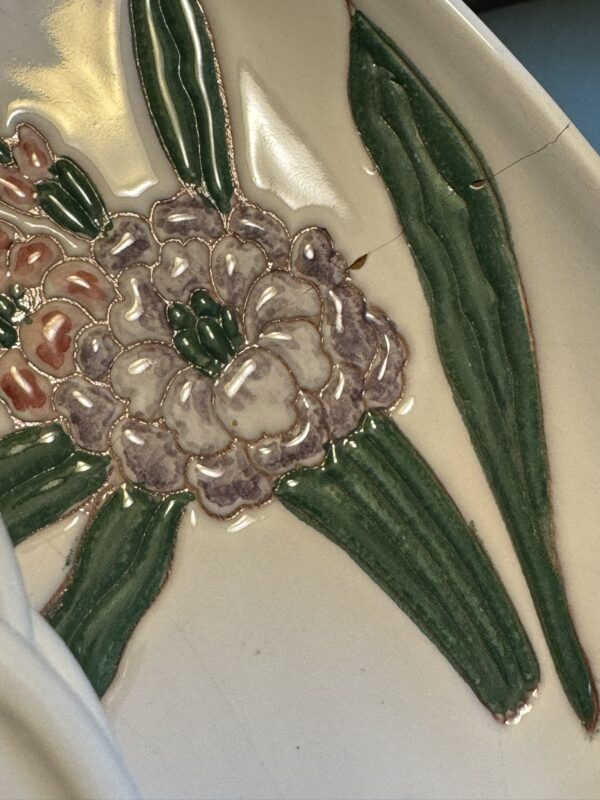 Close-up of a ceramic plate with an embossed flower design. The flower features pale purple petals, pink buds, and green leaves. A visible crack runs through the plate, highlighting its delicate condition. The surface is glossy and slightly reflective.
