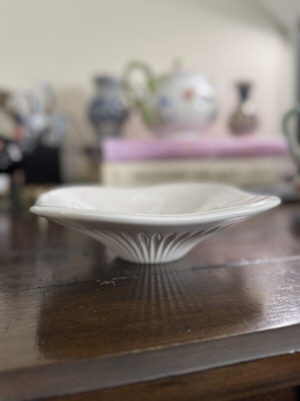 A white, shallow ceramic bowl with subtle embossed patterns sits on a dark wooden surface. In the softly blurred background, books and various decorative vases and mugs are visible, adding a cozy, homey atmosphere to the scene.