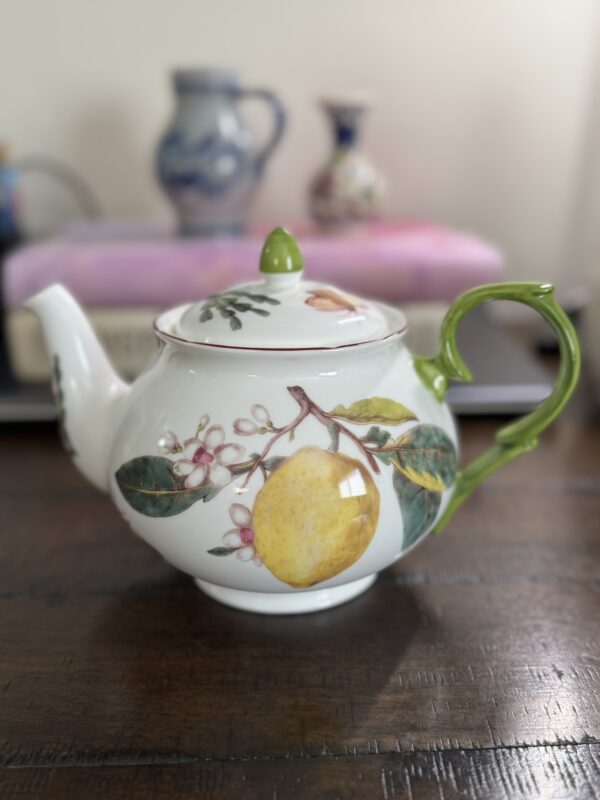 A porcelain teapot with a painted yellow lemon, green leaves, and pink blossoms on its side. The handle is green; the lid has a green knob. The teapot sits on a dark wooden surface with blurred books and vases in the background.