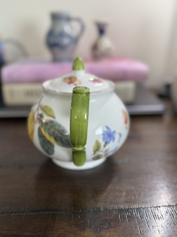 A round porcelain teapot with a green handle and knob sits on a dark wooden table. The teapot features colorful floral and fruit designs. In the blurred background, a stack of books, a pink cloth, and ceramic vases are visible.