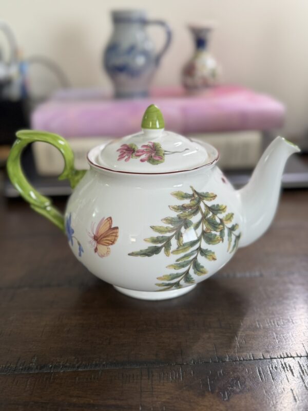 A white porcelain teapot with a green handle and lid knob, decorated with painted butterflies, flowers, and leafy branches, sits on a dark wooden table. In the blurred background, two decorative vases and a stack of books are visible.