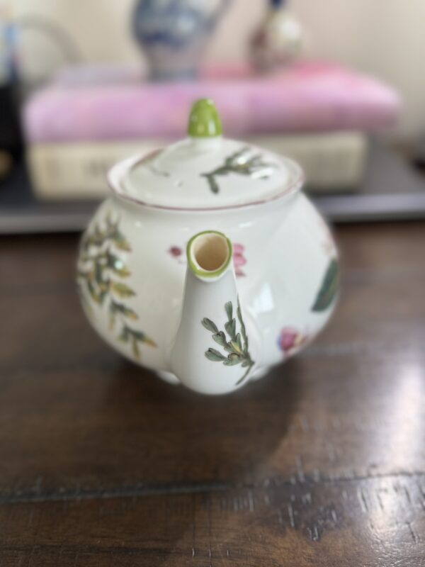 A white porcelain teapot with a floral design, a green knob on the lid, and pink accents is placed on a wooden table. The spout faces forward. In the blurred background, books and decorative objects are partially visible.