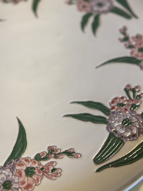 A close-up of a ceramic plate with hand-painted floral designs along the rim. The flowers are pink and purple with green leaves, arranged in clusters against a cream-colored background. The image focuses on the delicate detailing of the artwork.