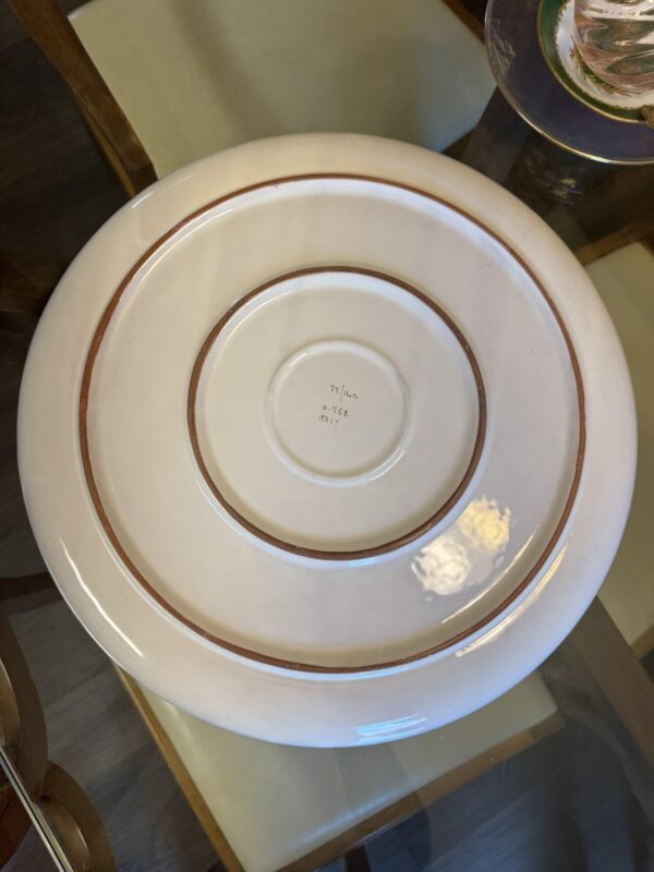 A large, round, white ceramic plate is upside down on a glass table. The base features two unglazed rings and handwritten text that reads Italy, Pasta, 3174. The table and chairs are visible around the plate.