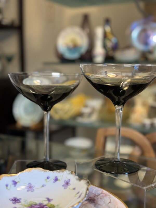 Two coupe glasses filled with a dark beverage sit on a table. In the foreground, there is a decorative plate with a floral pattern. The background shows shelves with various porcelain plates and decorative items, slightly out of focus.