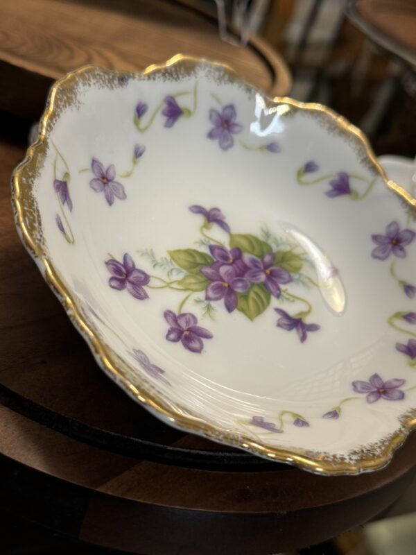 A small, scalloped-edged porcelain dish with a gold rim features a floral pattern of purple violets and green leaves. The dish rests on a dark wooden surface, and the intricate design gives it an elegant, vintage appearance.
