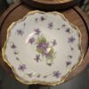 A porcelain plate with scalloped, gold-rimmed edges features a painted pattern of purple flowers with green leaves. The plate sits on a wooden surface, and its delicate floral design is prominent in the center and around the rim.