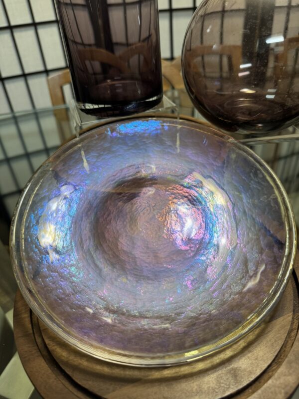 A glass bowl with an iridescent, pearl-like surface reflecting blue, purple, and pink hues sits on a wooden plate. Behind it, two smoky purple glass vases and a glass tabletop are visible, with a black grid background and wooden chair also in view.