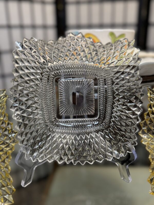 A clear, square, pressed glass plate with an intricate diamond pattern stands upright on a transparent display stand. The plate has a textured, geometric design radiating from the center and a small square indented area in the middle.