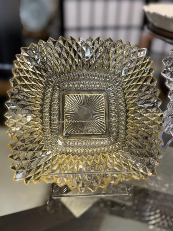 A clear square glass dish with intricate geometric diamond and starburst patterns, reflecting golden hues. The dish has sharply textured edges and is displayed upright on a glass surface, with a blurred background.