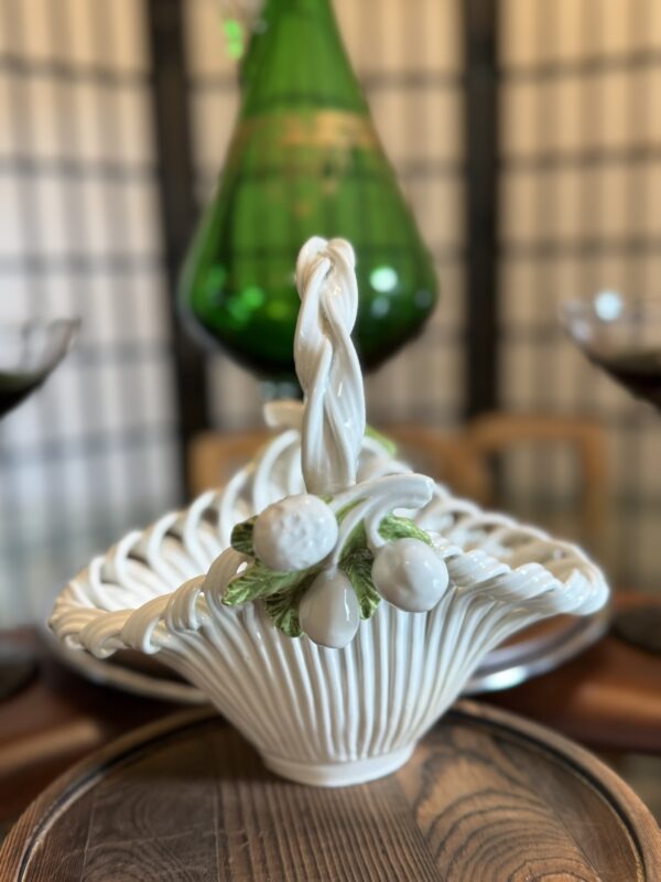 A white ceramic basket with a twisted handle and decorative white berries and green leaves sits on a wooden tray. In the blurred background, a tall green glass decanter and black goblets are visible against a folding screen and wooden chairs.