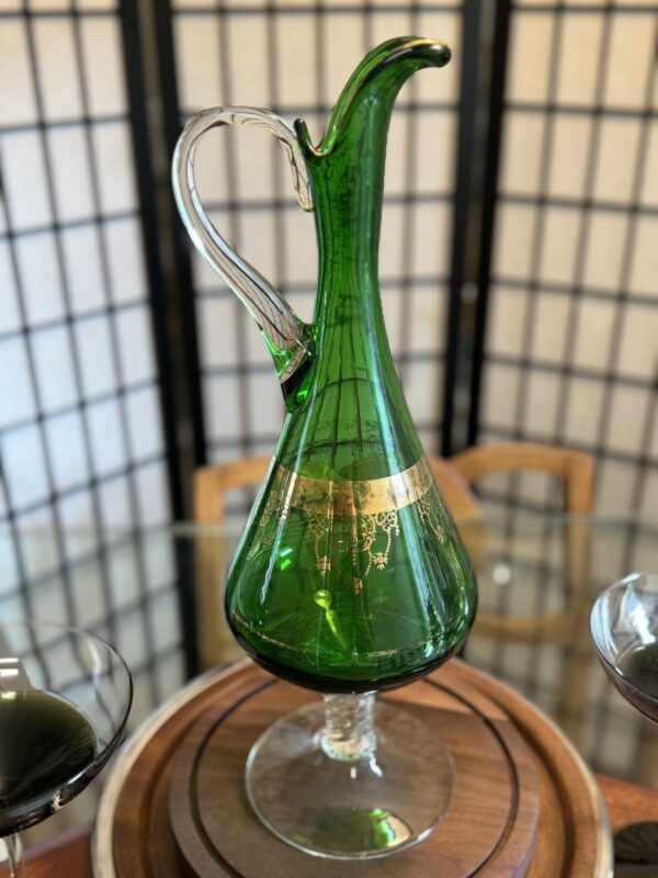 A tall, elegant green glass decanter with a gold decorative band around its widest part is displayed on a wooden tray. The decanter has a curved spout, a clear handle, and sits in front of a wooden screen with geometric patterns.