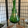 A tall, elegant green glass wine decanter with a clear handle and base sits on a wooden tray. The decanter has a gold decorative band around its body and is displayed in front of a folding screen and surrounded by glassware.